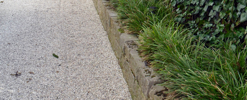 Gartenweg aus Quarzsplitt führt entlang einer Natursteinmauer aus Muschelkalkblöcken. Die Natursteinmauer ist mit Gräsern und Säulenkirschen bepflanzt.